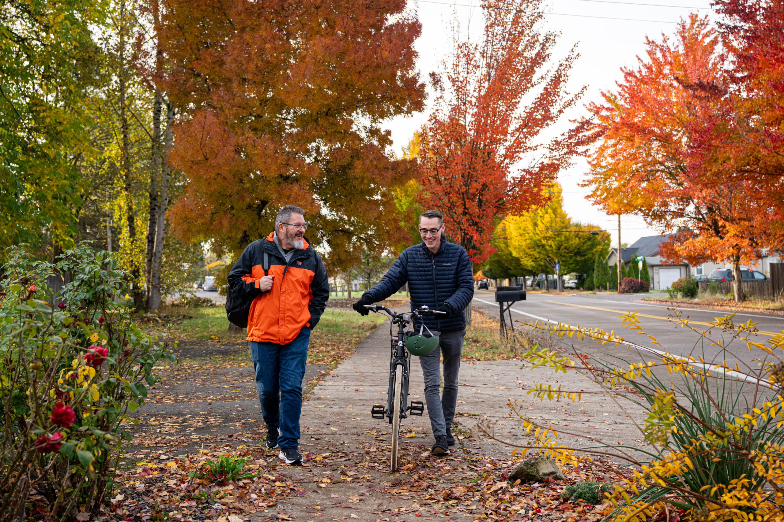 Two people walking down a neighborhood sidewalk in the fall, one with a bike and the other with a backpack.