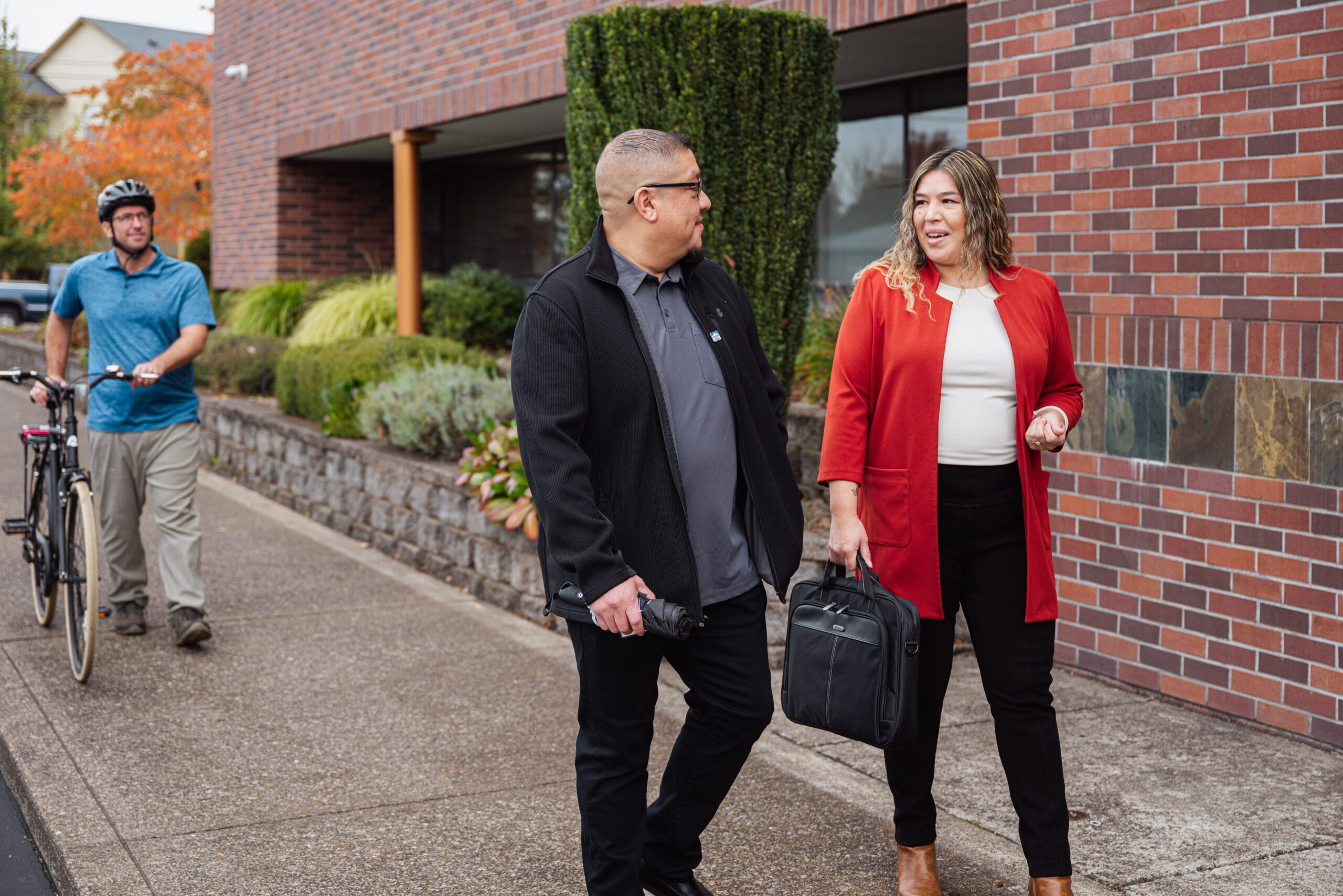 Three people walking along a brick building dressed in businesswear, one alongside a bicycle.