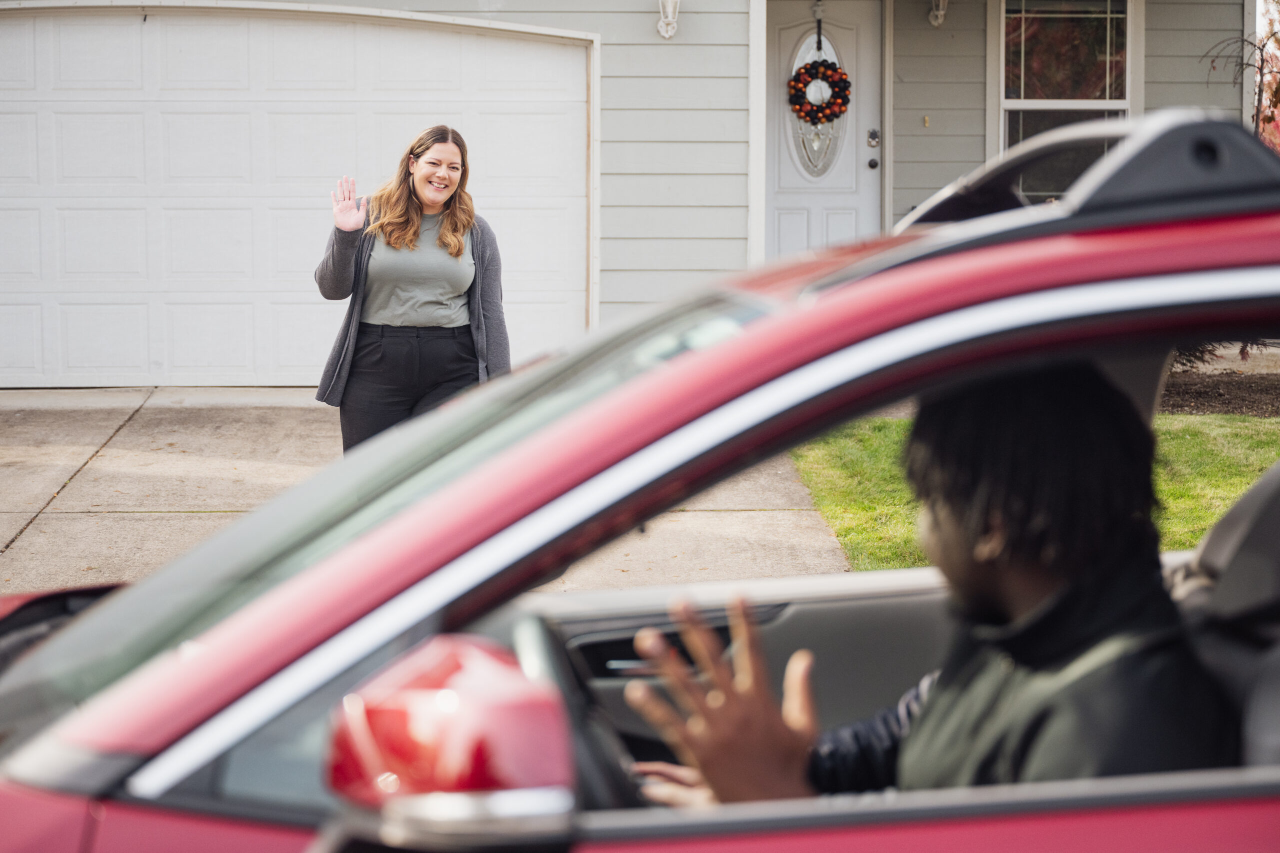 A person in a suburban driveway waving to the driver of a passing car.