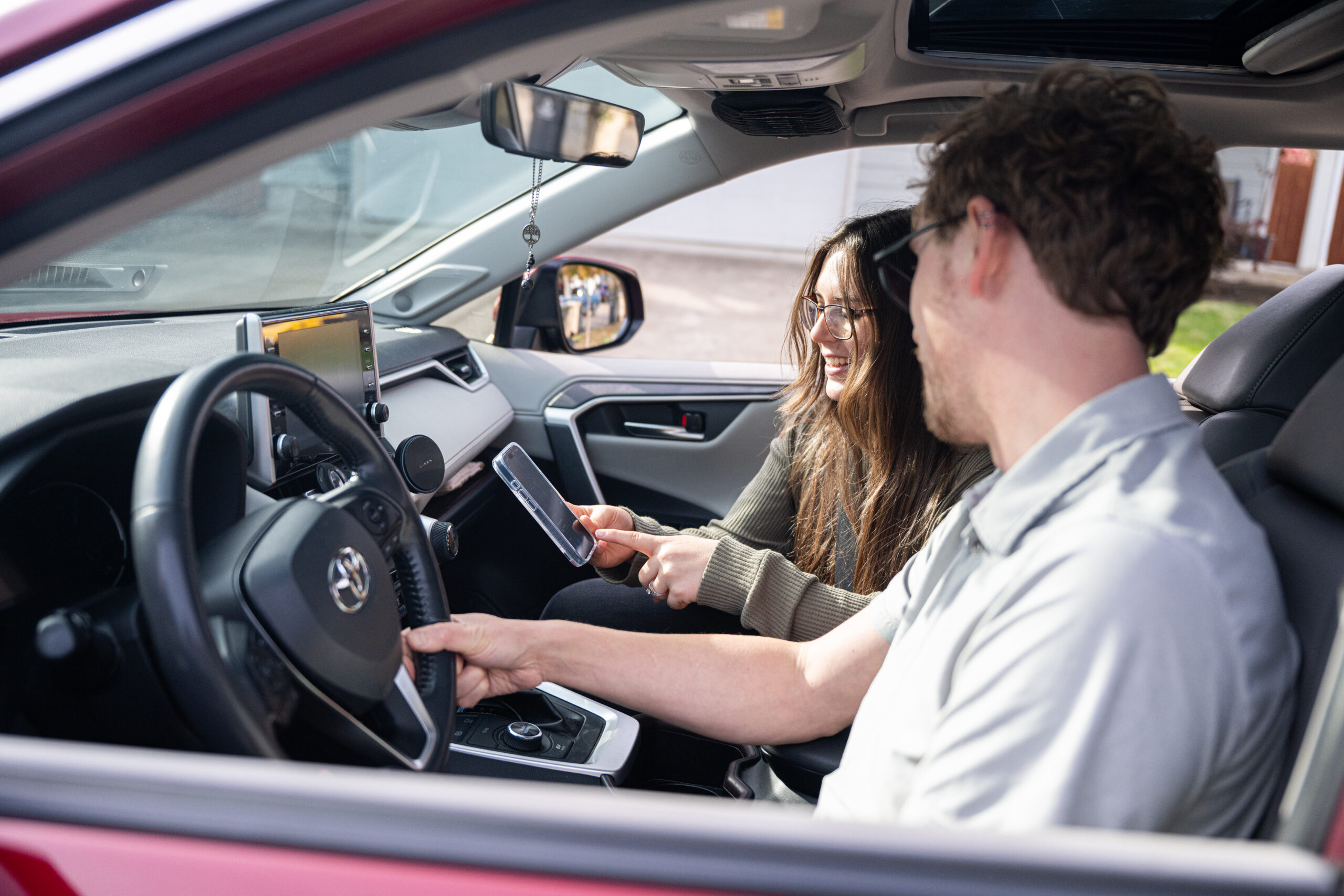 Two people in the front row of a parked car looking at a phone.