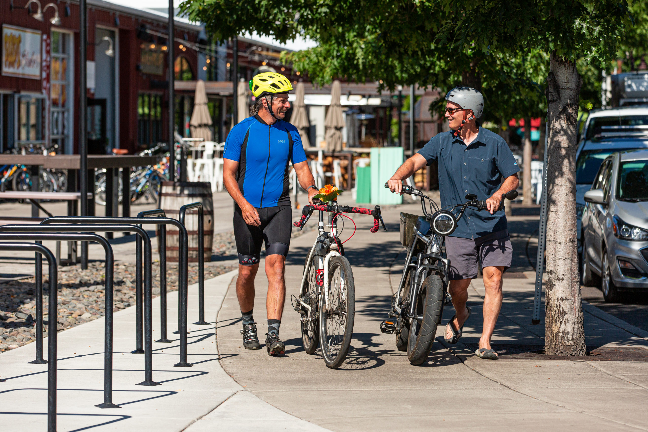 Two men walking bikes side by side on a city sidewalk.