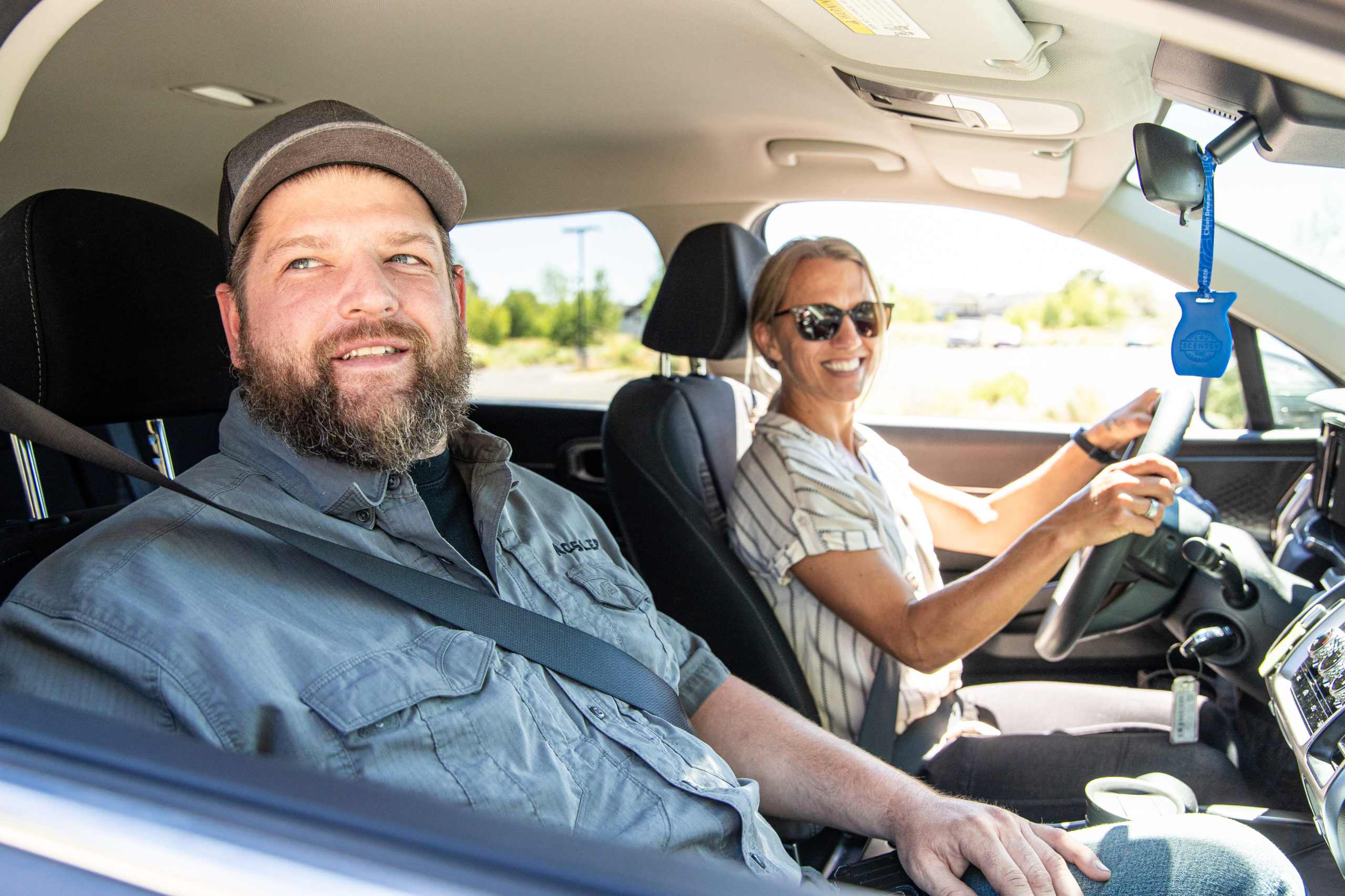 Two people in the front row of a car smiling at the passenger side window.