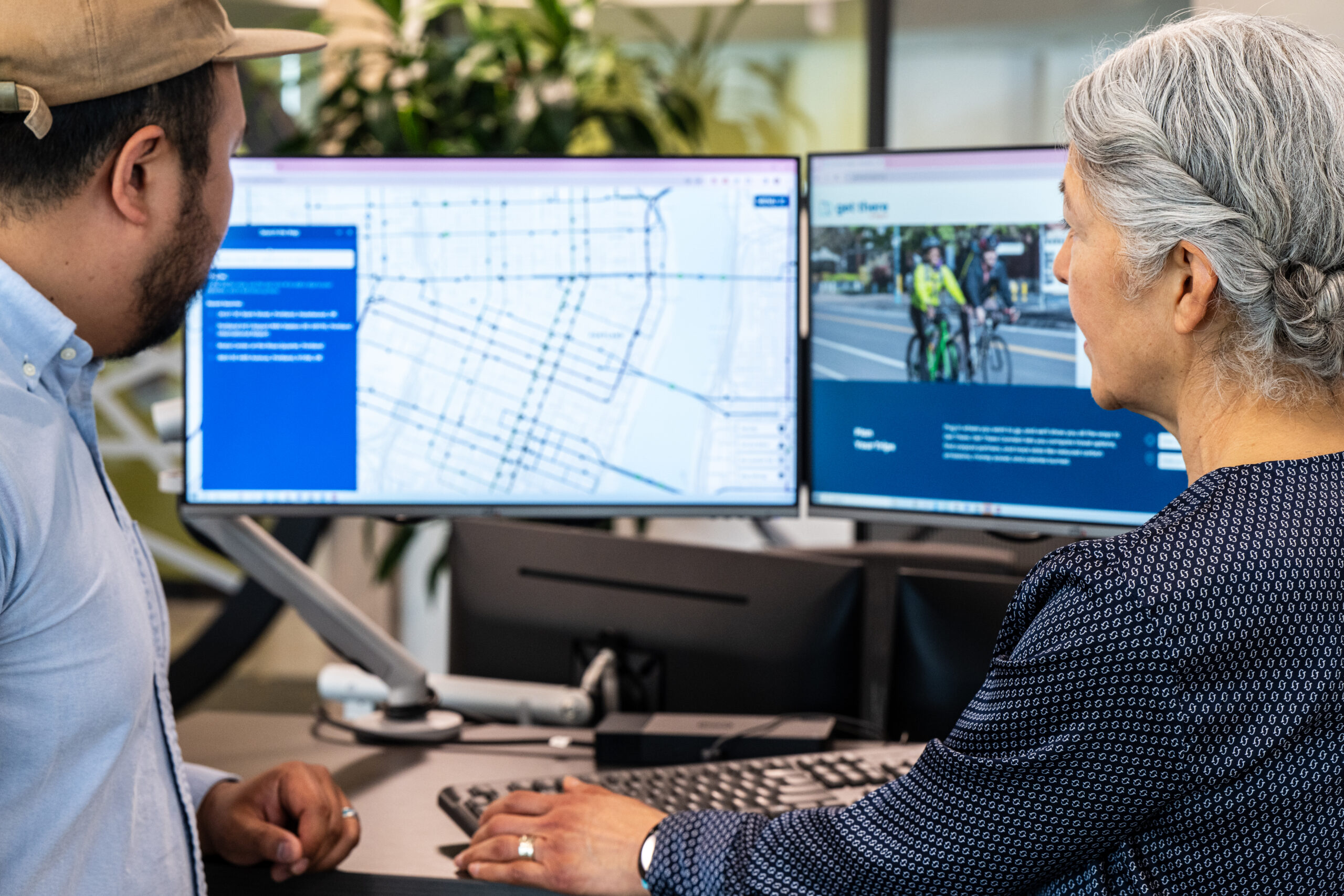 Two people standing and looking at double monitors showing the Get There website.